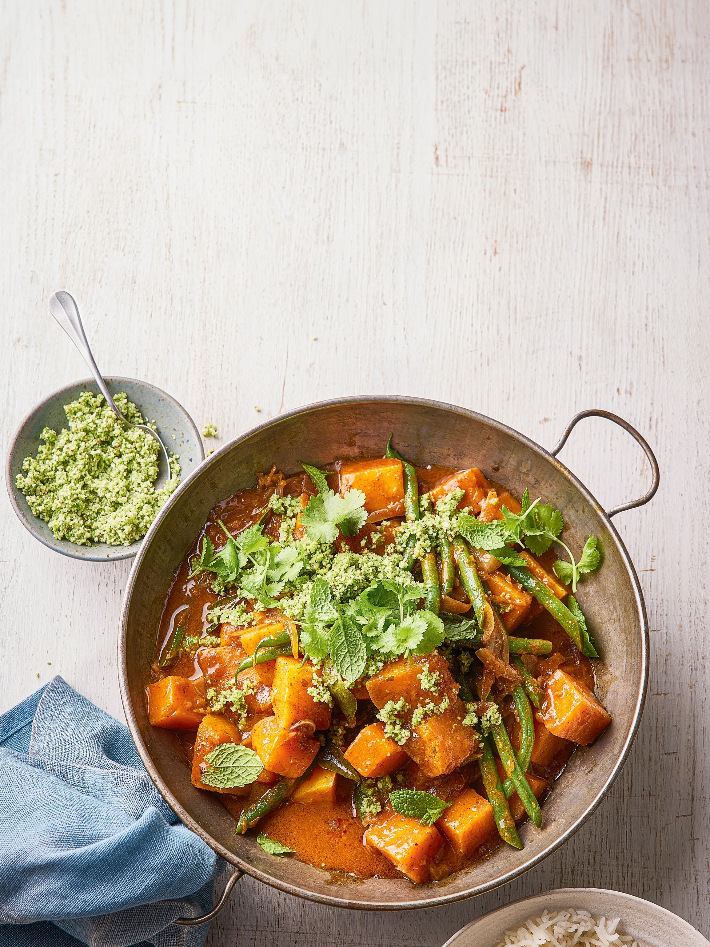 A vibrant dish featuring chunks of pumpkin and green beans garnished with fresh herbs and a sprinkle of green topping, served alongside a bowl of rice.